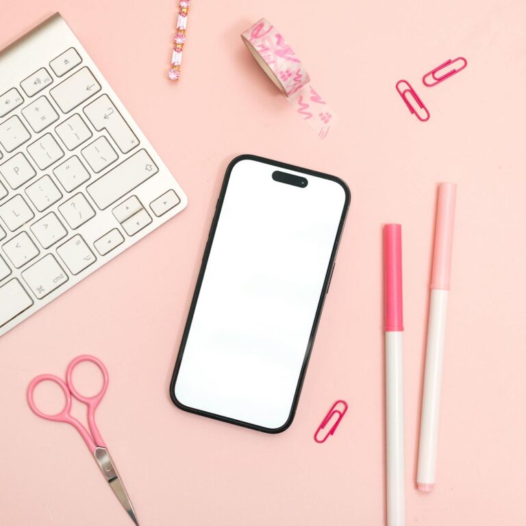 Flat lay of a pink themed office desk setup with an iPhone, keyboard, and stationery.
